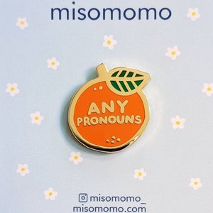 May include: A gold enamel pin with an orange and green design. The orange has the text "ANY PRONOUNS" on it. The pin is on a blue background with white flowers.