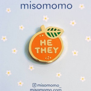 May include: Orange enamel pin with a green leaf and the words "HE THEY" in white letters. The pin is on a blue background with white flowers.
