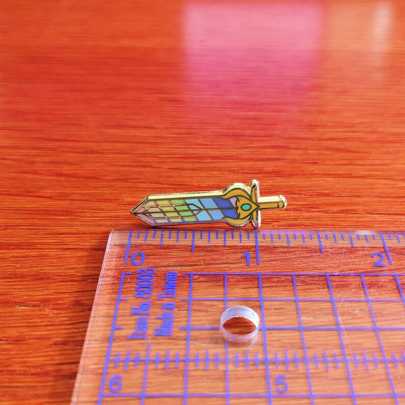 May include: A gold-colored enamel pin in the shape of a sword, featuring a colorful stained-glass design in shades of blue, green, and yellow. The pin is approximately 1.5 inches long and has a detailed hilt with a green gem.