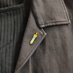 May include: A gold-colored sword-shaped enamel pin with a rainbow gradient blade and a green gem on the hilt. The pin is affixed to a dark gray blazer, creating a striking contrast. The pin is approximately 1 inch in length.