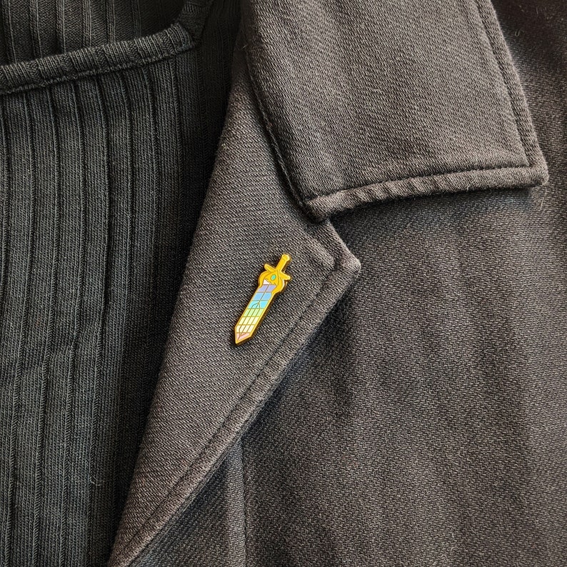 May include: A gold-colored sword-shaped enamel pin with a rainbow gradient blade and a green gem on the hilt. The pin is affixed to a dark gray blazer, creating a striking contrast. The pin is approximately 1 inch in length.