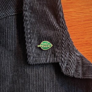 May include: A gold pin with a green leaf design. The leaf has the words "They Them" written on it.