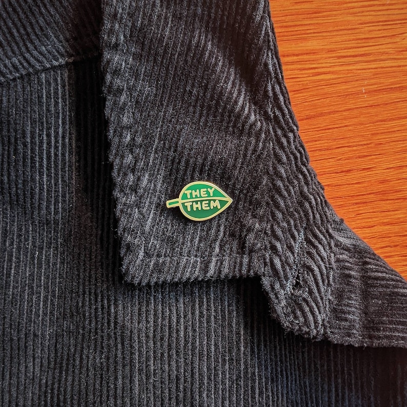 May include: A gold pin with a green leaf design. The leaf has the words "They Them" written on it.