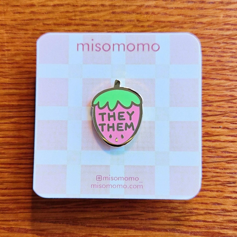 Puede incluir: Un pin de esmalte de fresa rosa con las palabras "They Them" en texto negro. El pin est&aacute; sobre un fondo a cuadros blanco y rosa.
