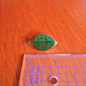 May include: A gold-colored pin shaped like a green leaf with the words "SHE HER" written in gold letters.