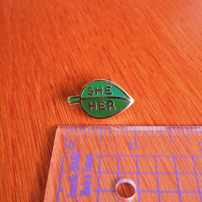 May include: A gold-colored pin shaped like a green leaf with the words "SHE HER" written in gold letters.