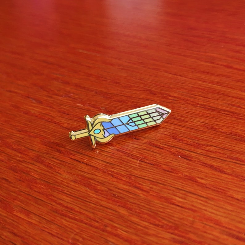 May include: A gold-colored sword pin with a colorful, stained-glass design on the blade. The pin features a detailed hilt and a geometric pattern of blue, green, and purple hues. The pin is a decorative accessory.