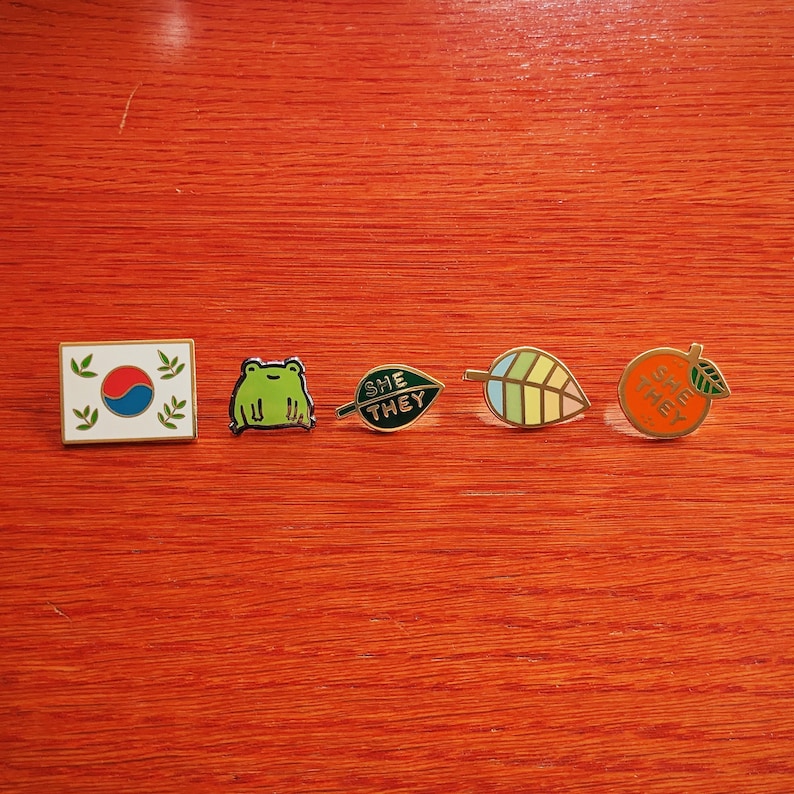 May include: Five enamel pins with different designs. The pins are on a red wooden surface. The pins are a green frog, a green leaf with the words "SHE THEY" on it, a green and yellow leaf, an orange with the words "SHE THEY" on it, and a white and red flag with green leaves on either side.