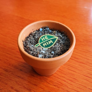 Puede incluir: Una maceta de terracota llena de tierra marr&oacute;n oscura y un pin de hoja verde con las palabras "HE HIM" en letras doradas.