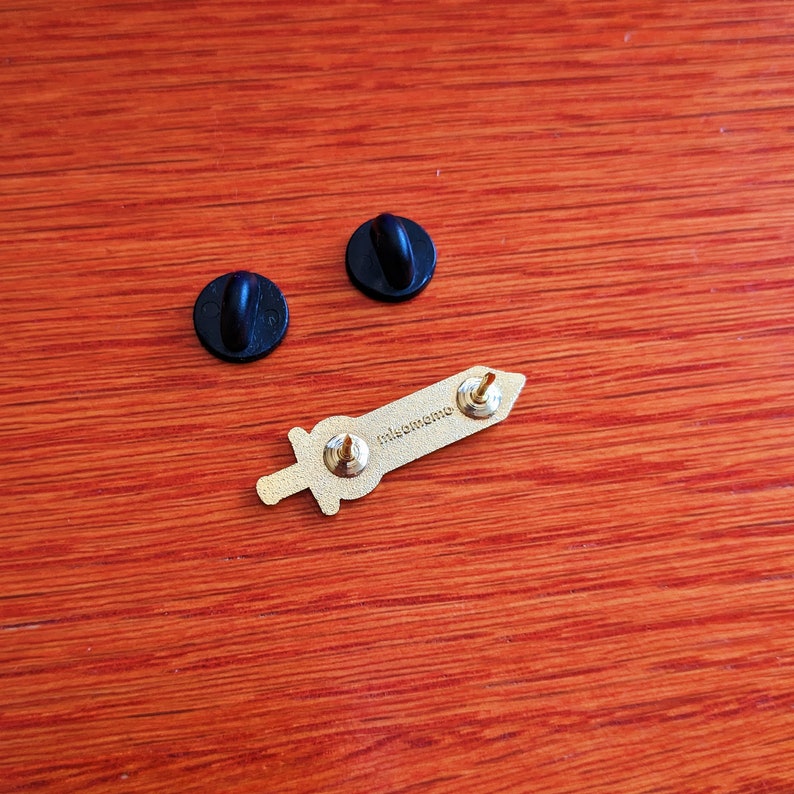 May include: A gold-colored sword-shaped enamel pin with the text "misomeme" on the back. Two black pin backs are also visible. The pin is on a wooden surface.