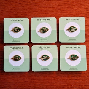 May include: Six green and white cards with the words "misomomo" printed on them. Each card features a gold pin with a green leaf design and text on the leaf. The text on the pins reads "She Her", "They Them", "He Him", "She They", "Any Pronouns", and "He They".