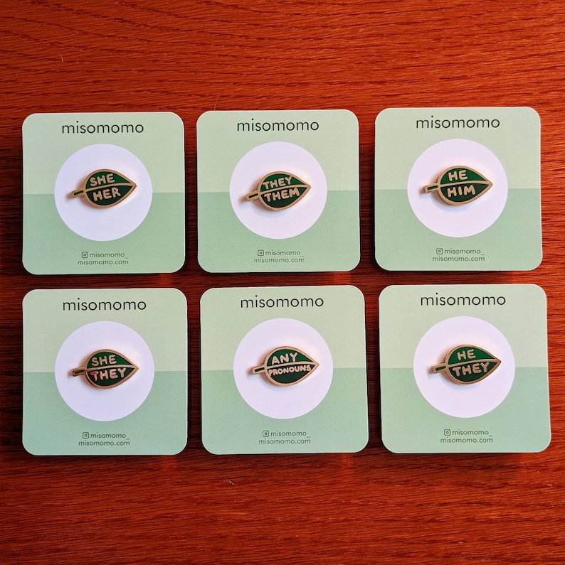 May include: Six green and white cards with the words "misomomo" printed on them. Each card features a gold pin with a green leaf design and text on the leaf. The text on the pins reads "She Her", "They Them", "He Him", "She They", "Any Pronouns", and "He They".