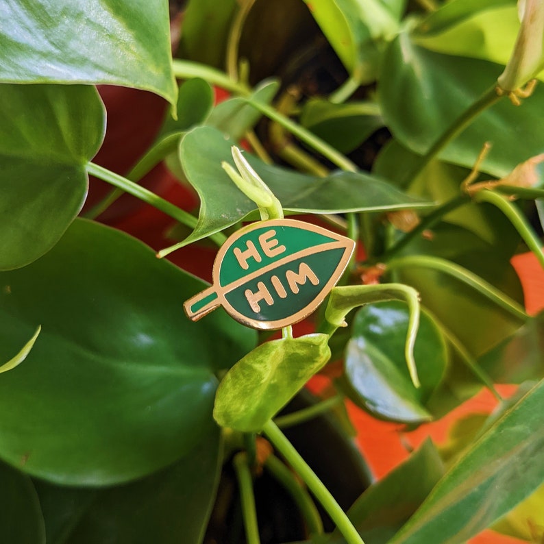 Puede incluir: Un pin de color dorado con forma de hoja verde con las palabras "HE HIM" escritas en &eacute;l.