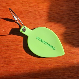 May include: A green leaf-shaped keychain with a silver metal ring and the word "misomomo" printed in green.