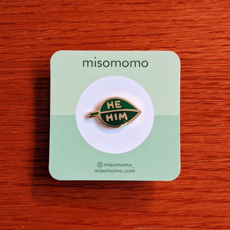 Puede incluir: Un pin de esmalte en forma de hoja verde con las palabras "HE HIM" en letras doradas. El pin est&aacute; en una tarjeta blanca con el texto "misomomo" y el nombre de usuario de las redes sociales "@misomomo_" y el sitio web "misomomo.com".