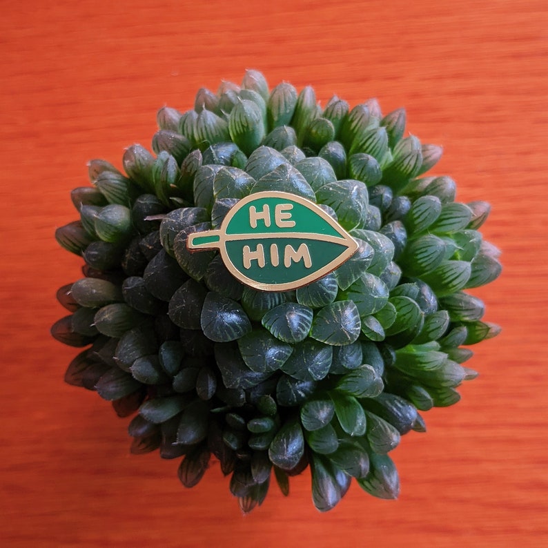 Puede incluir: Un pin de esmalte en forma de hoja verde con las palabras "HE HIM" en letras doradas.