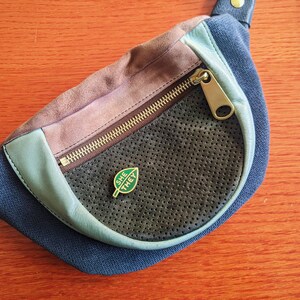 May include: A blue and brown fanny pack with a zipper closure and a green leaf pin with the words "SHE THEY" on it.