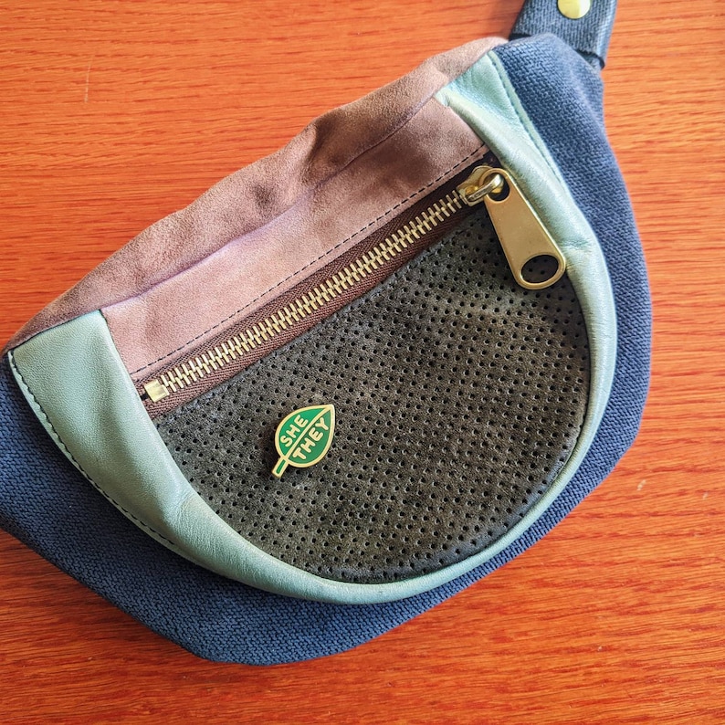 May include: A blue and brown fanny pack with a zipper closure and a green leaf pin with the words "SHE THEY" on it.