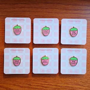 Puede incluir: Seis pines de esmalte en forma de fresa rosa con contornos dorados. Cada pin presenta un conjunto de pronombres diferente: "She Her", "They Them", "He Him", "She They", "Any Pronouns" y "He They".