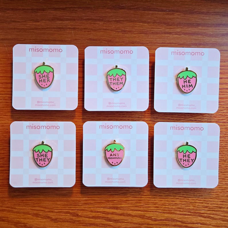 Puede incluir: Seis pines de esmalte en forma de fresa rosa con contornos dorados. Cada pin presenta un conjunto de pronombres diferente: "She Her", "They Them", "He Him", "She They", "Any Pronouns" y "He They".