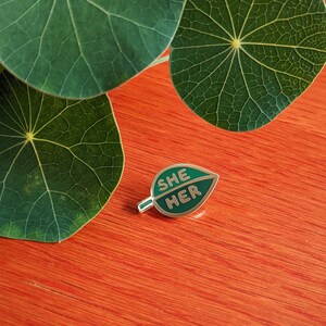 May include: A green leaf-shaped enamel pin with the words "SHE HER" in gold lettering.