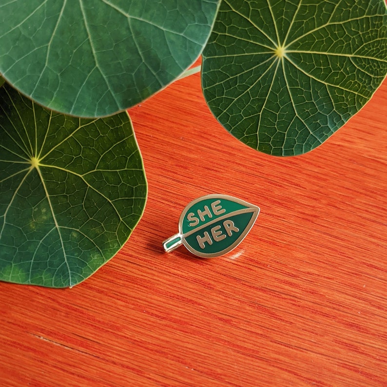 May include: A green leaf-shaped enamel pin with the words "SHE HER" in gold lettering.