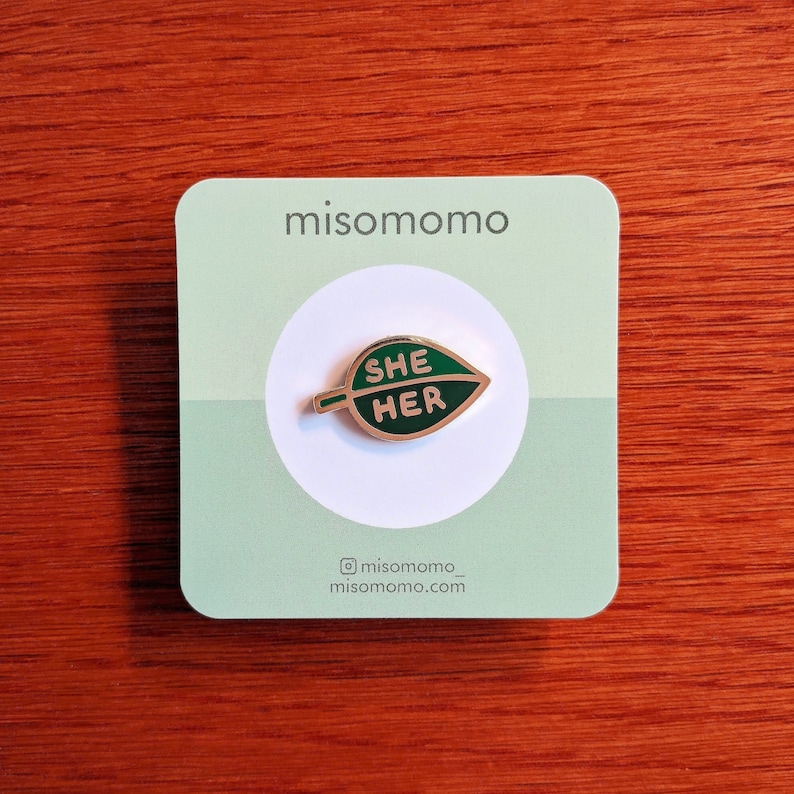 May include: A green leaf-shaped enamel pin with the words "SHE HER" in white lettering. The pin is on a white card with the text "misomomo" and the social media handle "@misomomo_" and website "misomomo.com".