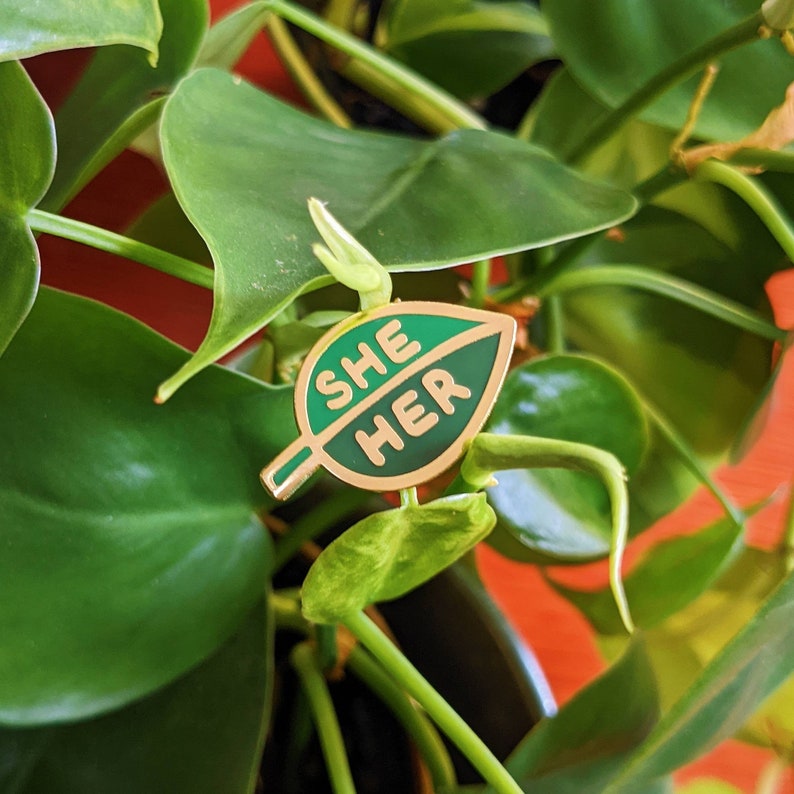 May include: A green leaf-shaped enamel pin with the words "SHE HER" in gold lettering.