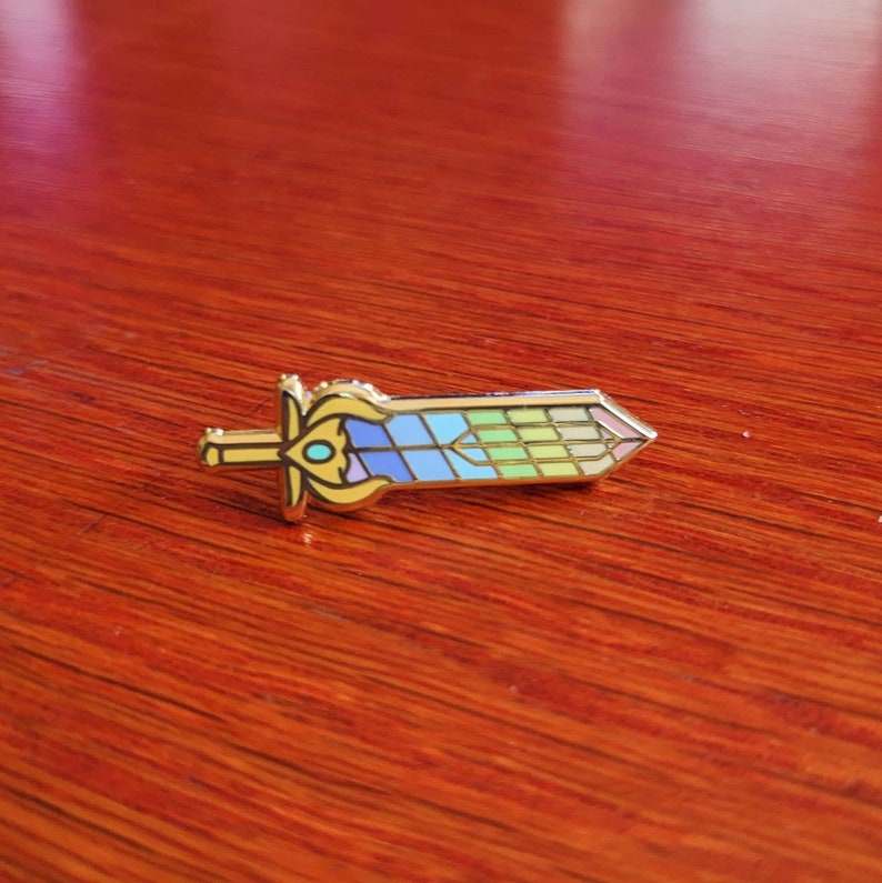 May include: A gold-colored sword-shaped enamel pin with a colorful stained-glass design. The pin features a detailed hilt and a blade with blue, green, and yellow segments. The pin is set against a wooden surface.