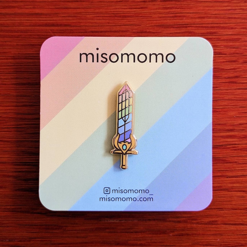May include: A gold-colored sword pin with a colorful, stained-glass design. The pin is on a pastel-colored card with the text "misomomo" and the Instagram handle "misomomo_". The sword has a blue, green, and purple color scheme.