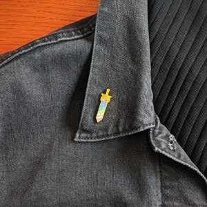May include: A colorful enamel pin in the shape of a sword, featuring a gold hilt and a blade with a rainbow gradient. The pin is attached to a dark denim jacket.