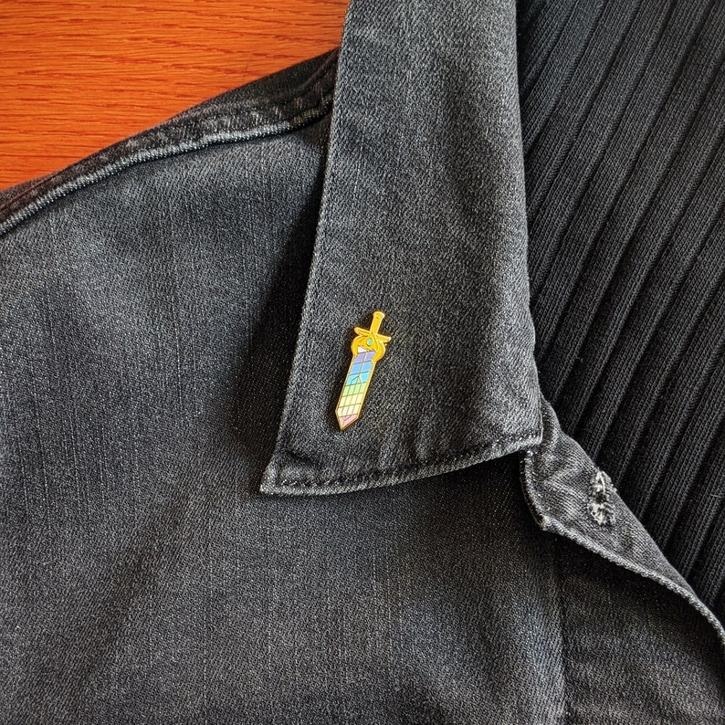 May include: A colorful enamel pin in the shape of a sword, featuring a gold hilt and a blade with a rainbow gradient. The pin is attached to a dark denim jacket.