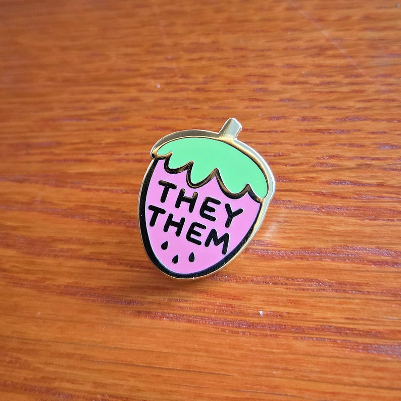 Puede incluir: Un pin de esmalte rosa y verde con forma de fresa con el texto "THEY THEM" en su interior.