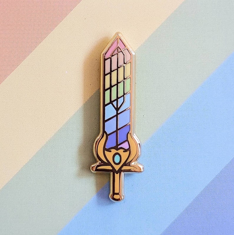 May include: A gold-colored enamel pin in the shape of a sword. The sword blade features a rainbow design with various colors. The hilt has a gold-colored design with a blue gem in the center. The pin is set against a pastel rainbow background.