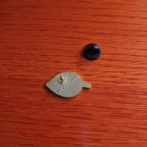 May include: Gold-colored leaf-shaped enamel pin with the text "misomomo" on the back. The pin has a black rubber backing.