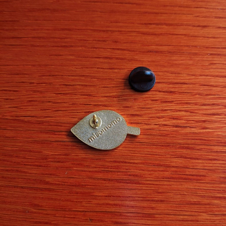May include: Gold-colored leaf-shaped enamel pin with the text "misomomo" on the back. The pin has a black rubber backing.