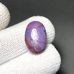 May include: An oval-shaped cabochon gemstone featuring a polished purple surface with visible reddish-brown inclusions and natural veining patterns. The stone has a smooth, reflective finish and is displayed against a neutral, textured background.
