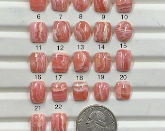May include: Twenty-two polished rhodochrosite cabochons in various shapes, including squares and ovals, are arranged in rows. Each stone displays unique pink and white banded patterns. A United States quarter coin is placed next to the stones for scale.