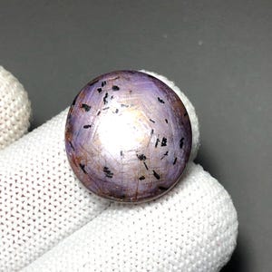 May include: A round, cabochon-cut gemstone with a polished, light purple surface. The stone features a distinct six-rayed star effect centered on its dome, interspersed with numerous small, dark, needle-like inclusions scattered throughout the body.