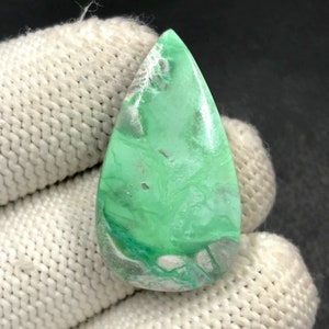 Puede incluir: Una gema pulida en forma de lágrima. Presenta un color verde vibrante con patrones arremolinados blancos y grises. La piedra se sostiene contra una tela de punto blanca.