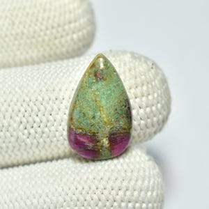Peut inclure: Une pierre précieuse polie en forme de goutte d'eau. La partie supérieure est vert moucheté, passant à un magenta vif à la base. La pierre est présentée sur un fond blanc texturé.