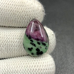 Puede incluir: Una piedra preciosa verde y rosa en forma de lágrima con manchas negras. La piedra preciosa está pulida y tiene una superficie lisa.