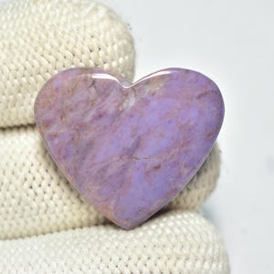 以下が含まれることがあります： ラベンダーとモーヴの色合いの、ハート型の研磨された宝石。 石は滑らかで光沢のある表面と自然な静脈を持っています。 ハートは白いテクスチャの背景に置かれています。