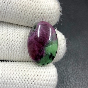 Puede incluir: Gema ovalada de rubí zoisita. La piedra pulida presenta un tono magenta intenso con inclusiones verdes y negras. La gema está sujeta por una mano enguantada de blanco sobre un fondo gris oscuro.