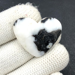 May include: A heart-shaped stone with a white base and black veining. The stone is held in a white knitted glove. The heart shape is smooth and polished, with the black veining creating a unique pattern across the surface.