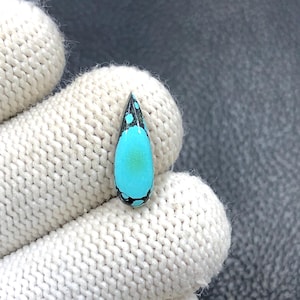 Op de afbeelding: Een druppelvormige edelsteen met een levendig turquoise centrum en zwart gespikkelde randen. De bovenkant van de steen is zwart met turquoise vlekken. De steen wordt vastgehouden door een witte gebreide handschoen. Deze edelsteen is ideaal voor het maken van sieraden.