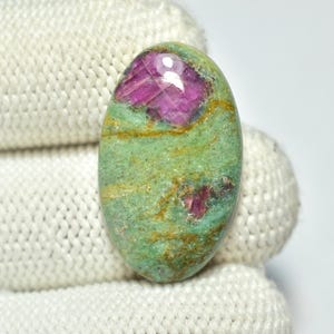 Peut inclure: Pierre précieuse ovale avec une inclusion de rubis rose vif dans une matrice verte. La pierre polie présente des motifs naturels et des veines dorées. Idéale pour la joaillerie ou la collection. Mesure environ 3 cm de long.