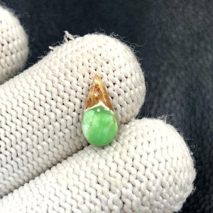 May include: A teardrop-shaped gemstone with a light green base, transitioning to a brown and gold top. A white band separates the colours. The gemstone is held in a white knitted glove. The gem is likely for jewellery making.