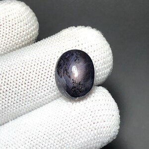 May include: An oval-shaped star sapphire gemstone with a deep dark blue and black colour palette. The stone features a distinct six-rayed star effect across its polished, cabochon-cut surface, displaying natural internal inclusions and dark mineral patterns.