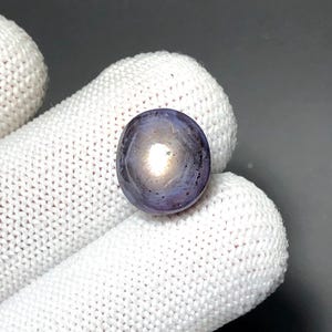 May include: A round, cabochon-cut star sapphire gemstone with a deep purple-blue hue. The stone features a distinct six-rayed asterism effect, appearing as a bright white star pattern centred on the smooth, polished surface of the translucent gem.